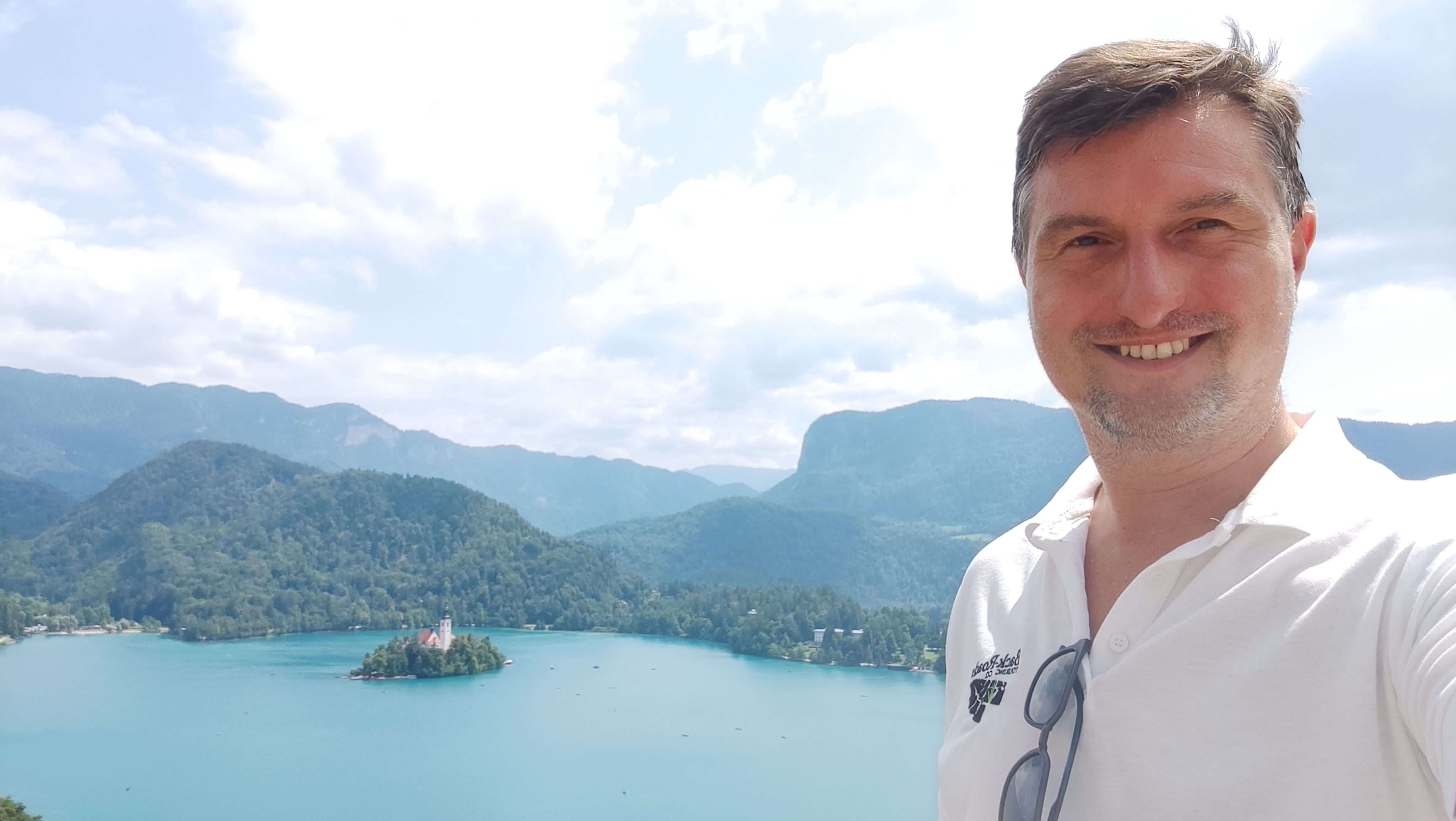 Selfie of man with salt and pepper hair wearing a white polo t-shirt in front of Lake Bled