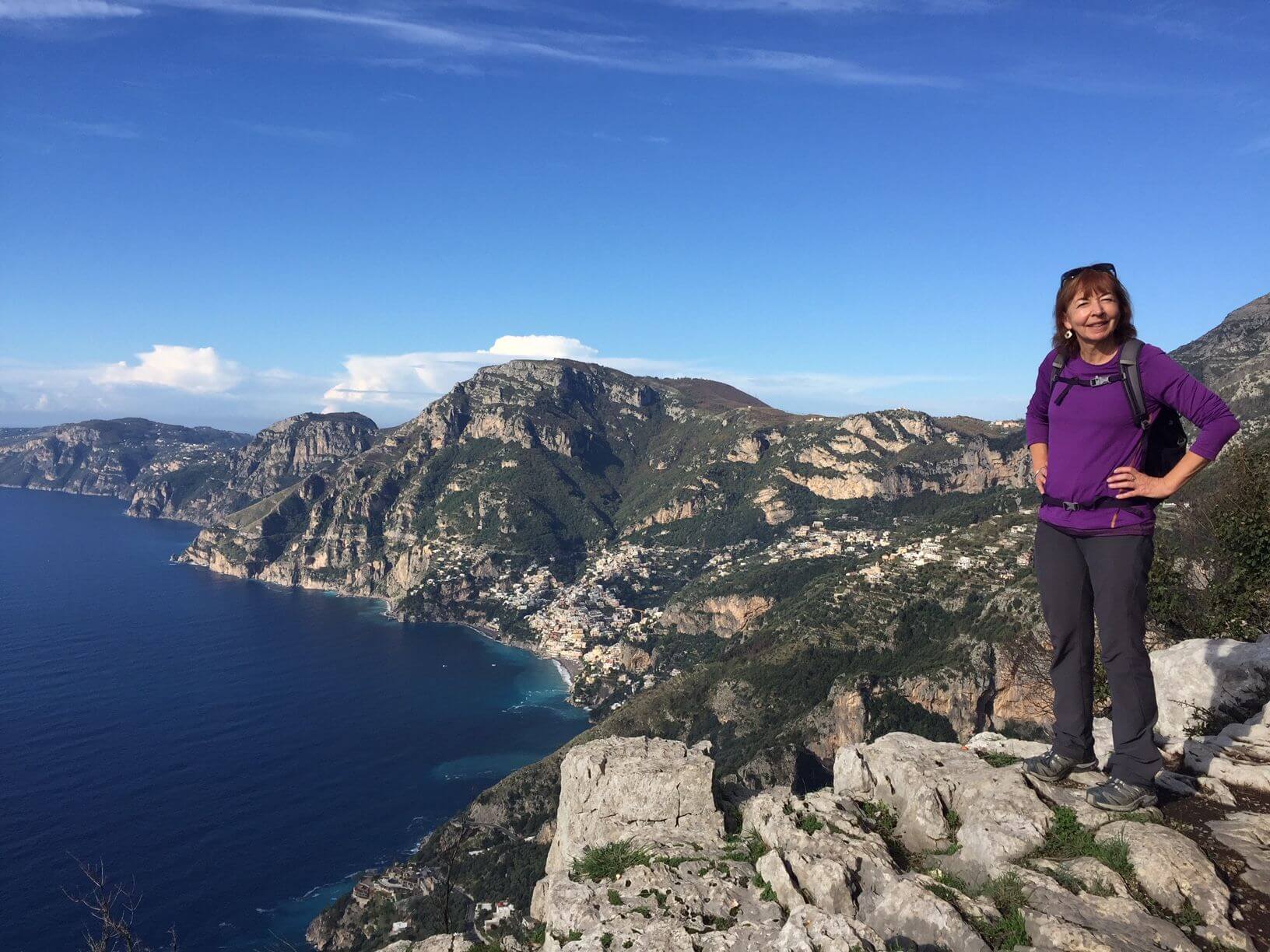 Female Tour Leader wearing purple long-sleeved top, standing on rocky hill with blue skies in background.