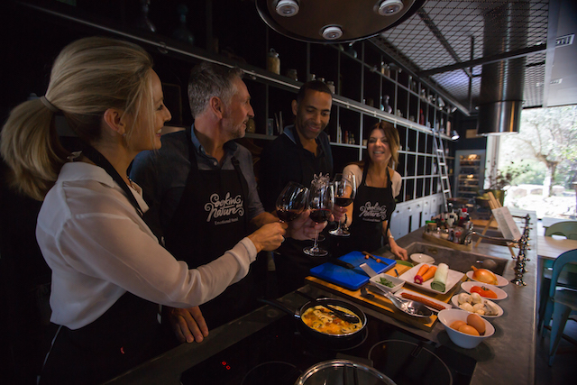 two pairs of couples wearing aprons and clinking wine glasses as they prepare a Portuguese meal in a professional kitchen