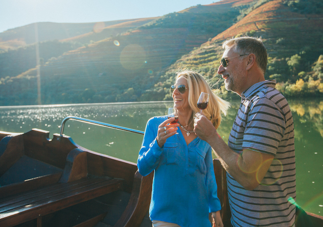 a middle aged couple wearing sunglasses on a boat in the sunny Douro Valley, Portugal, each with a glass of port