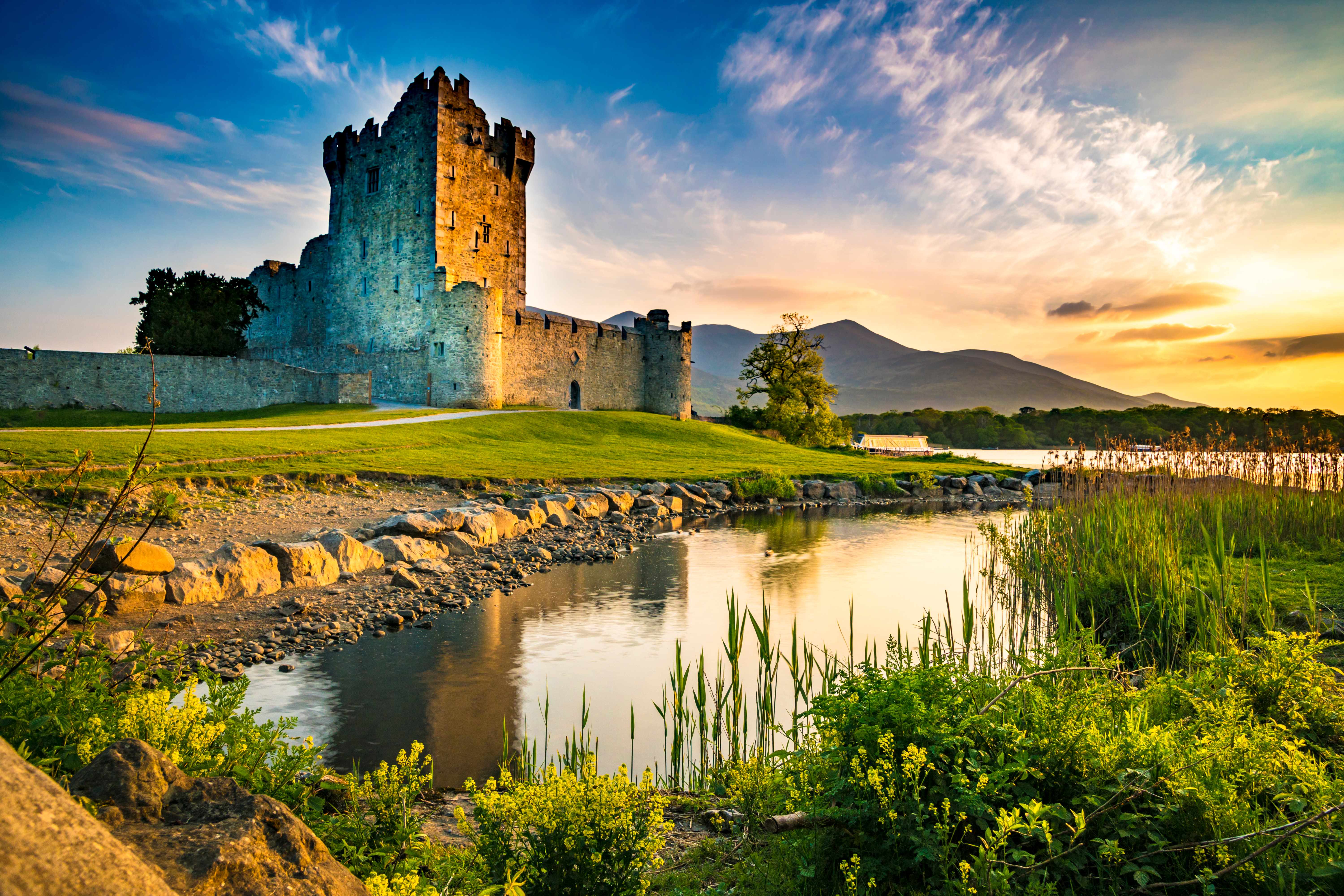 Stone fortress surrounded by lakes and greenery with mountains and sunrise in the background