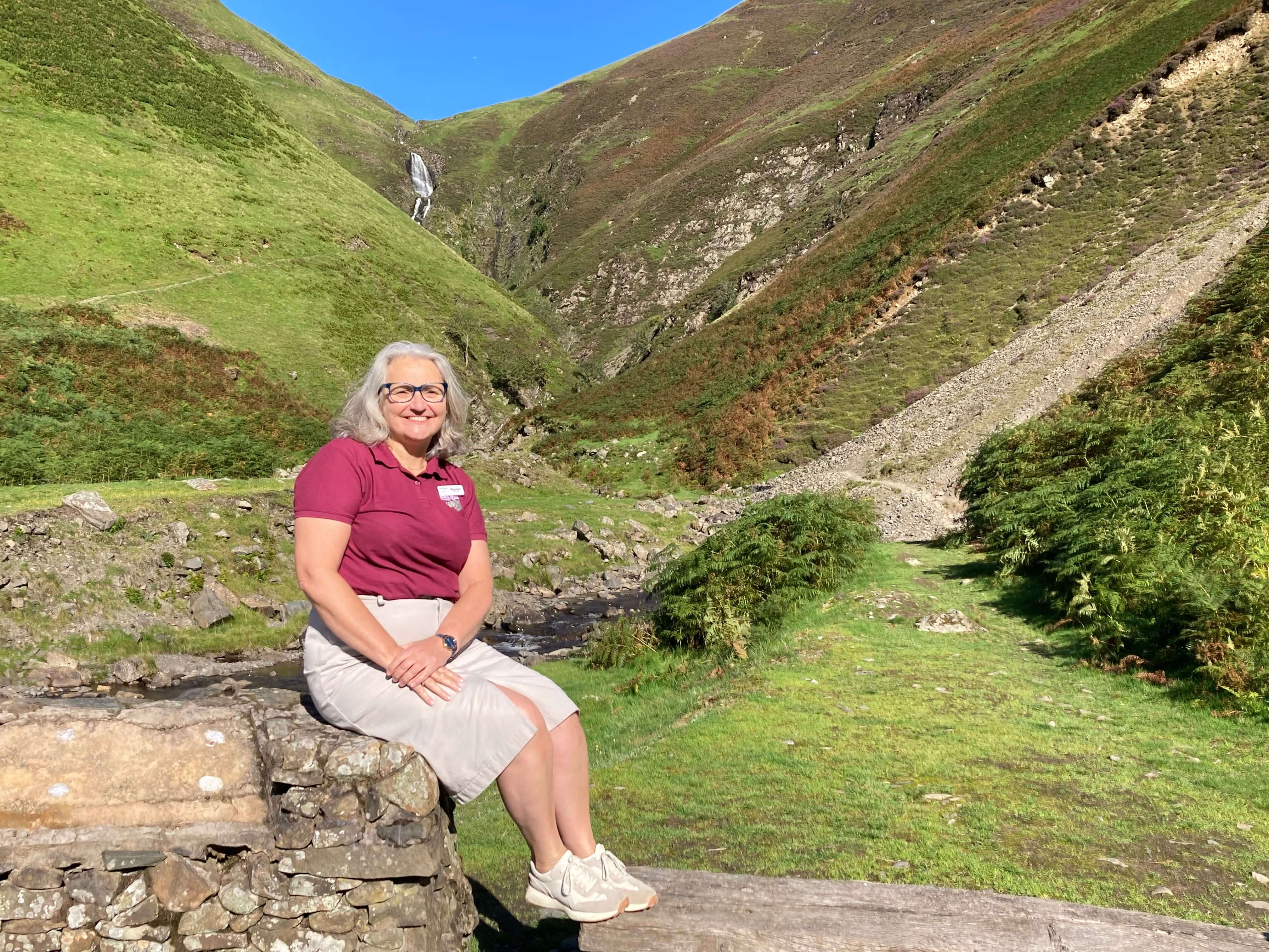 Mature woman smiling at camera whilst perched on a stone wall with rolling green hills in the background