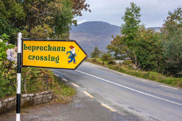 yellow road sign reading 'leprechaun crossing' with an empty road and mountain backdrop