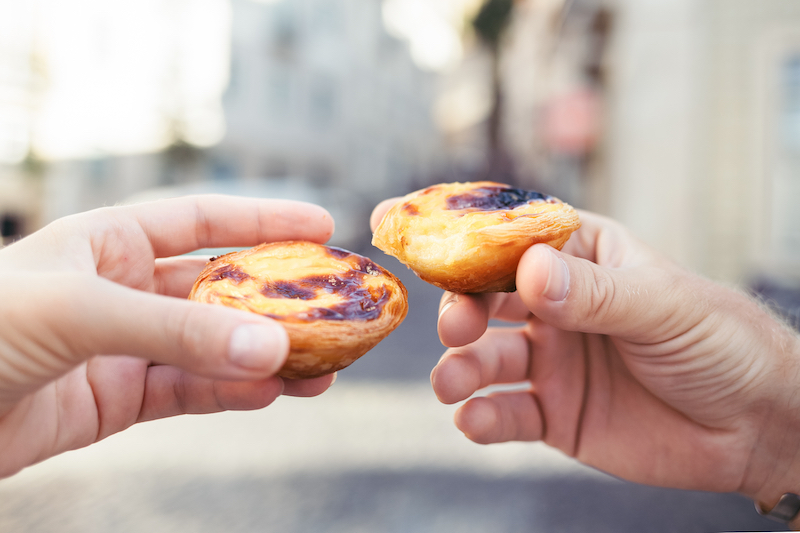 two hands holding Pastéis de nata, Portuguese egg tarts, with an out of focus Portugal street scene in the background