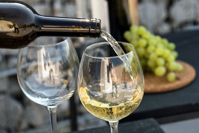 a pair of wine glasses with a bottle of white wine being poured with green grapes on a table in the background