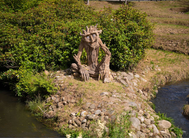 wooden statue of a cross-legged goblin sitting in a green area next to a river