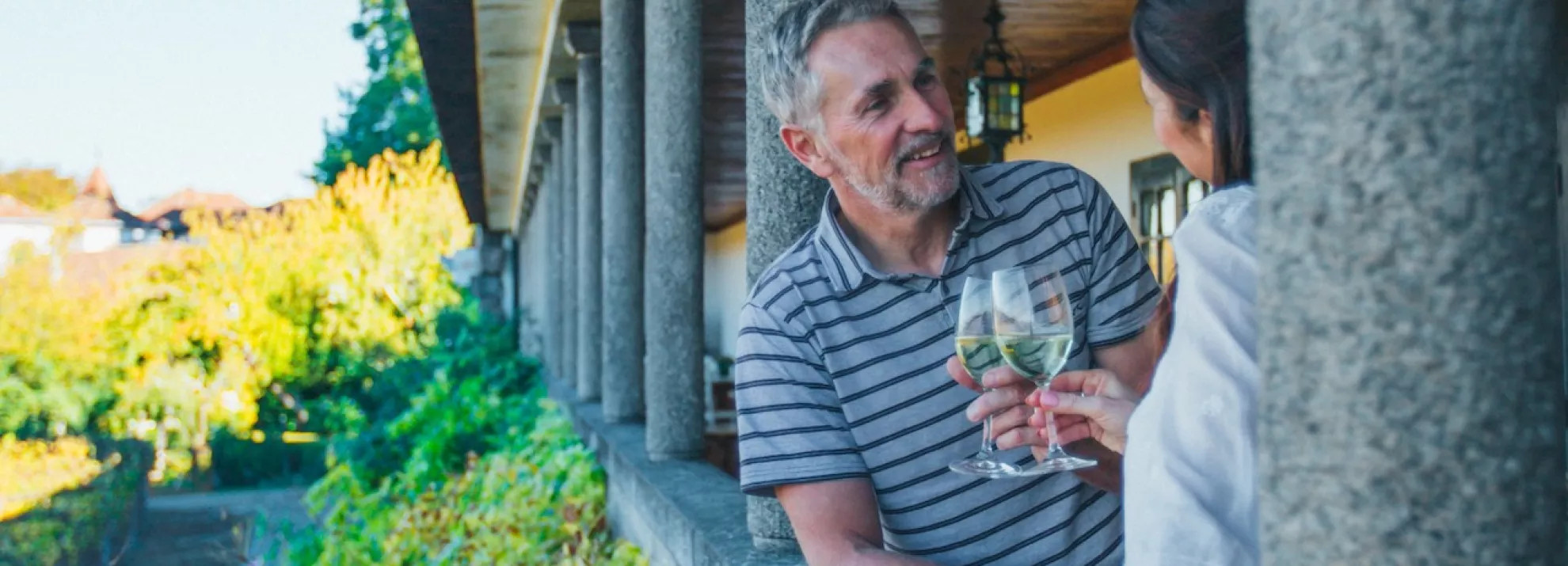 middle aged man and woman clinking glasses of white wine in a portico overlooking a leafy garden