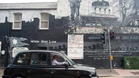 A black taxi cab driving past a mural in Belfast.