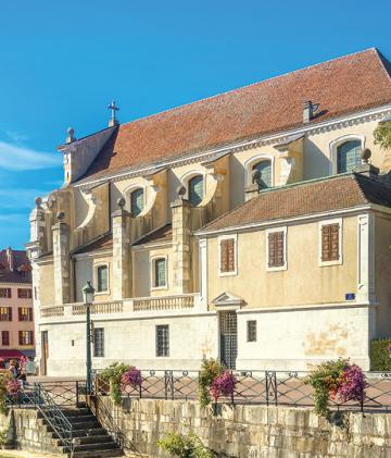 A large chateau stands on the coast of Lake Annecy, with some flowers in the foreground.